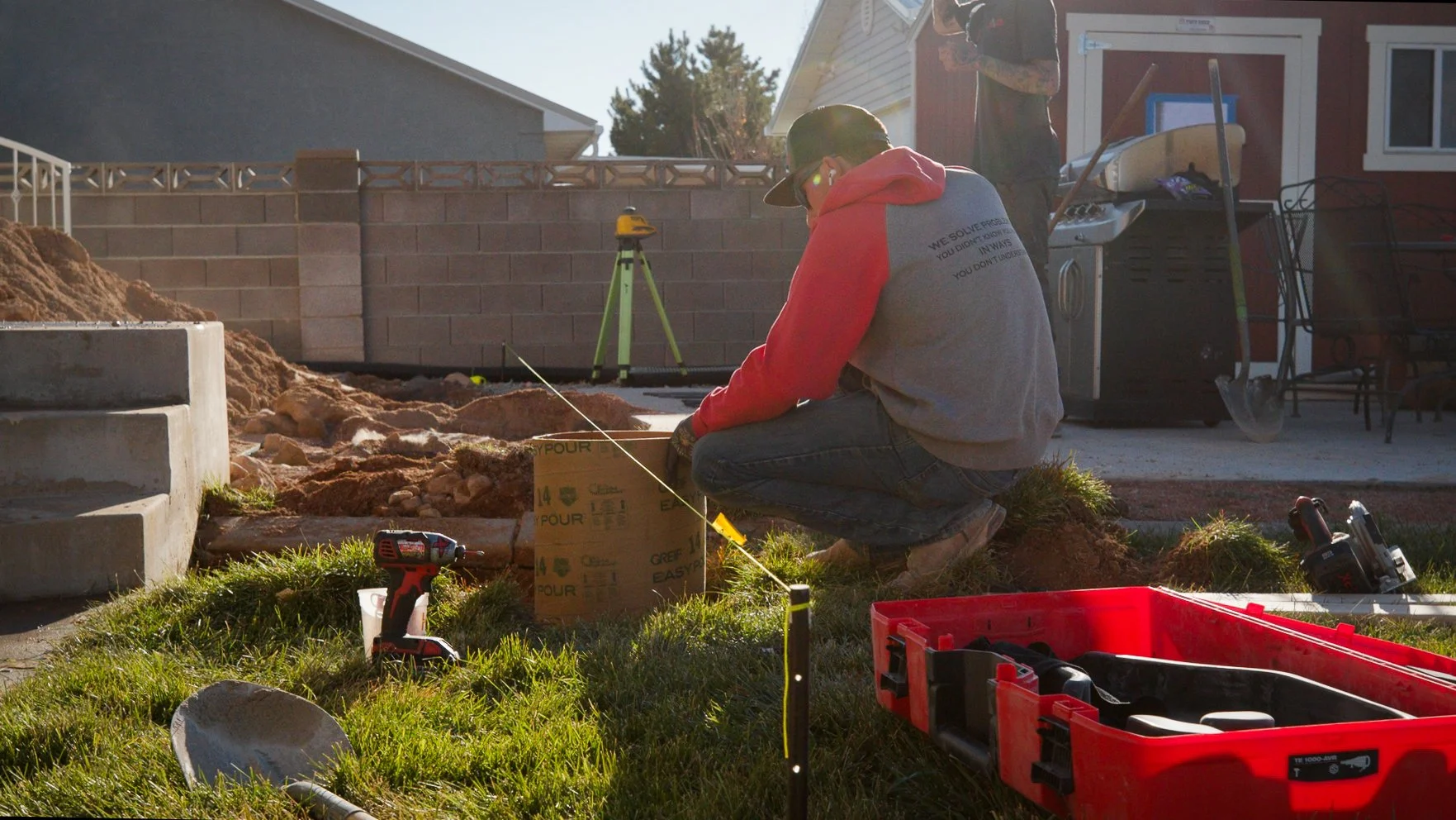 Person working on a landscaping project in a backyard, using tools and measuring tape, with a brick wall, patio, and house in the background.