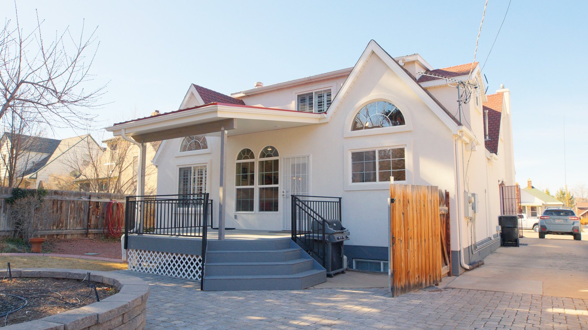 Backyard view of a white two-story house with large arched windows, a small porch with stairs, a black railing, a barbecue grill, and a fenced area, under a clear blue sky.