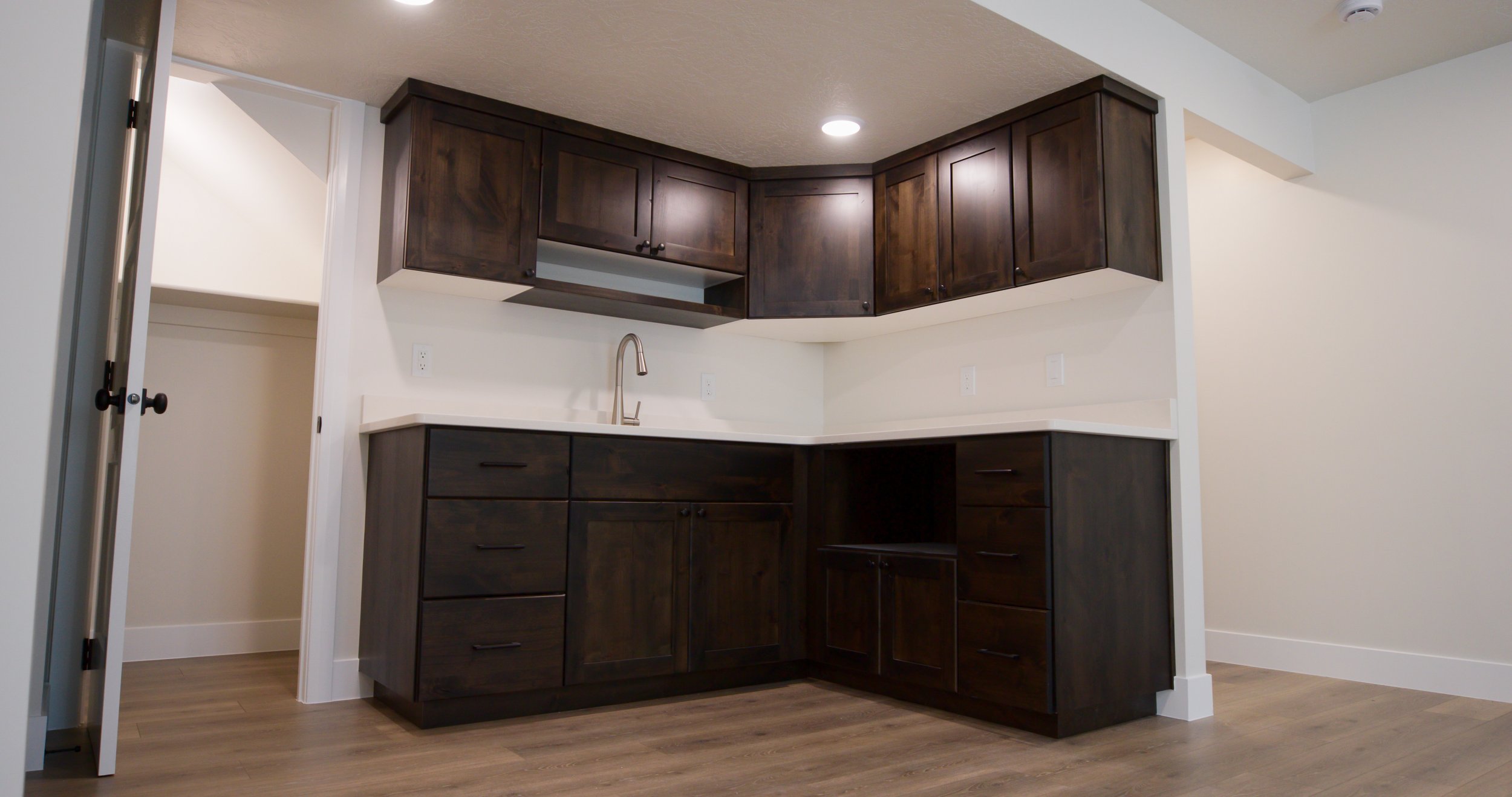 A modern kitchen with dark wood cabinets, a white backsplash, and a white countertop with a stainless steel faucet. The kitchen is in an open space with wood flooring and white walls.