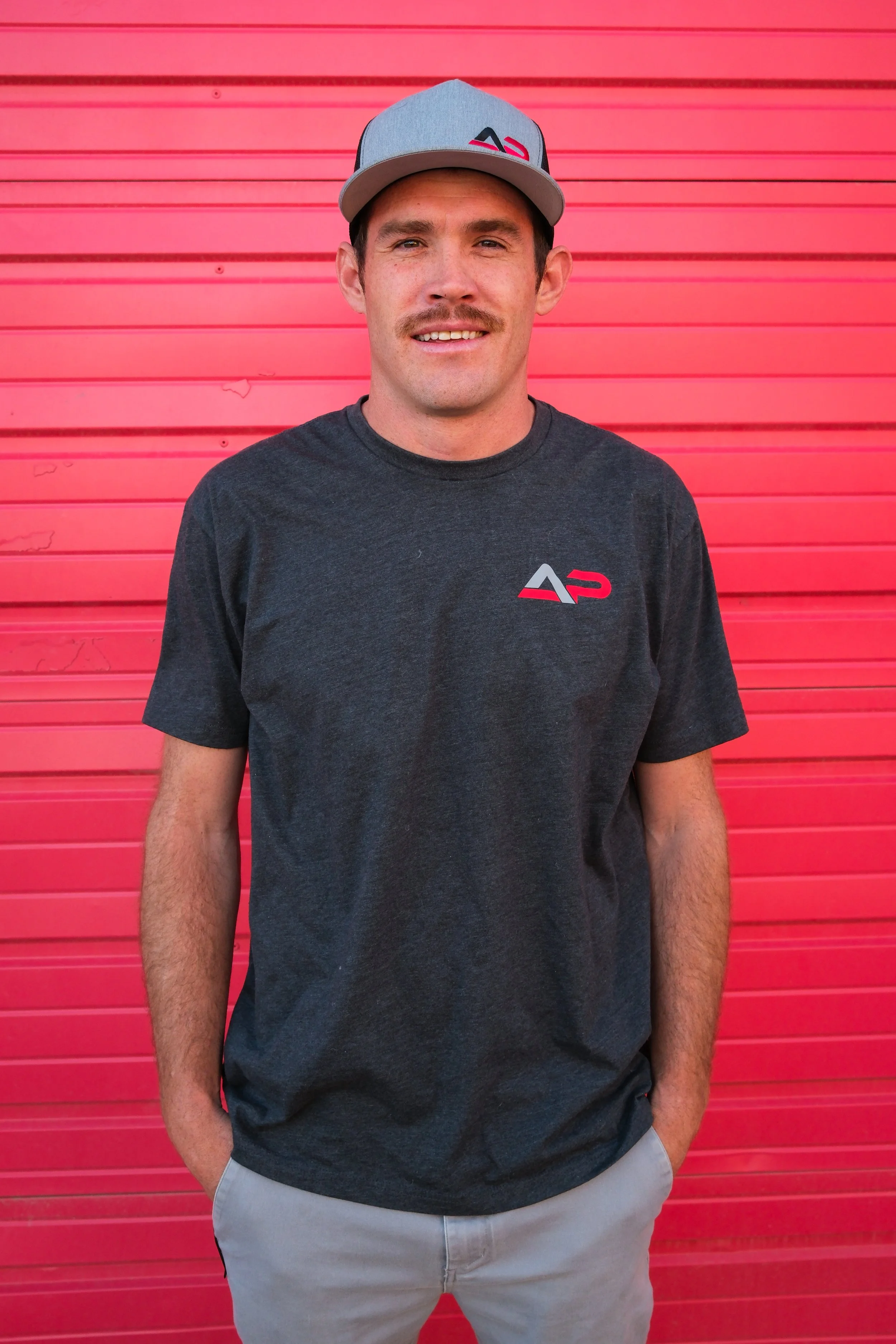 A man smiling, wearing a black t-shirt and a black cap with a logo, standing in front of a red wooden wall.