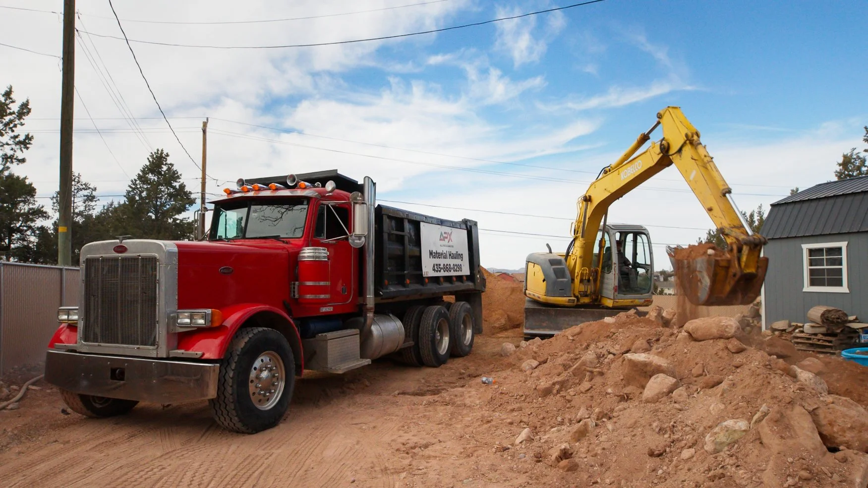 A red dump truck and a yellow excavator working on a construction site with dirt and rocks under a blue sky.