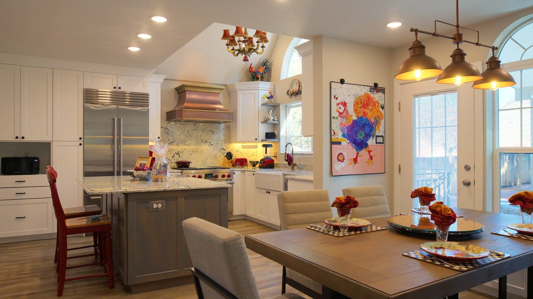 A kitchen and dining area with white cabinets, a granite countertop island, stainless steel refrigerator, and a stove with a copper range hood. There are red kitchen appliances, and a window with an arched top. The dining table has place settings wit