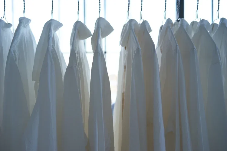 Rows of white laboratory coats hanging on hooks in front of a window with natural light.
