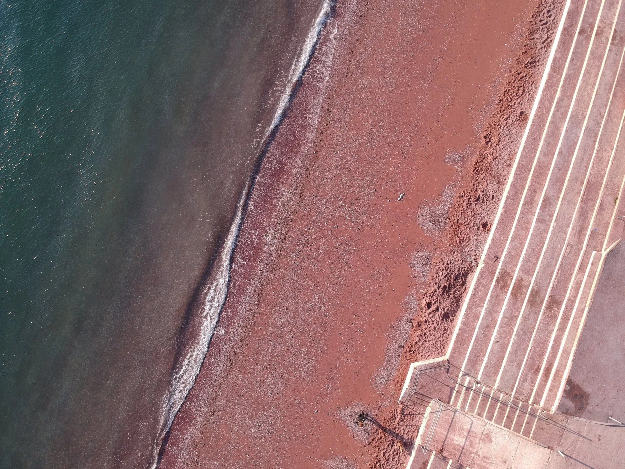 Aerial view of a pink beach with white lines marking parking spaces, and a few people walking along the shoreline with waves breaking.