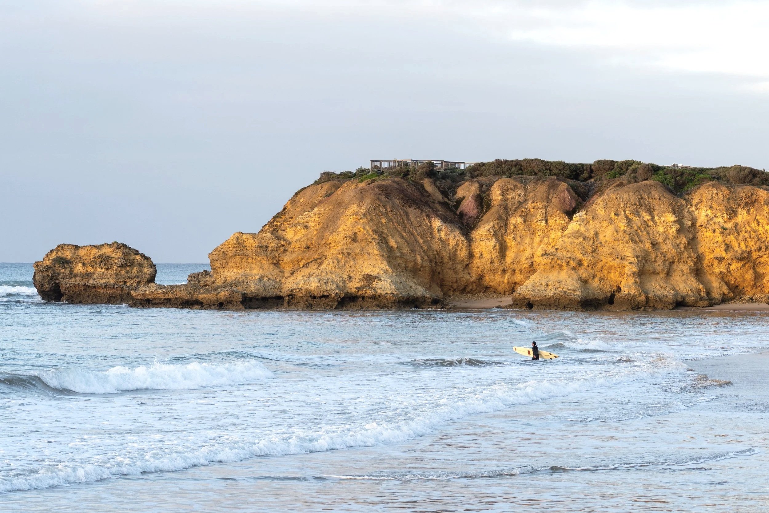 A surfer in a wetsuit holding a surfboard walking in the shallow ocean water near large rocky cliffs with a coastal building on top.