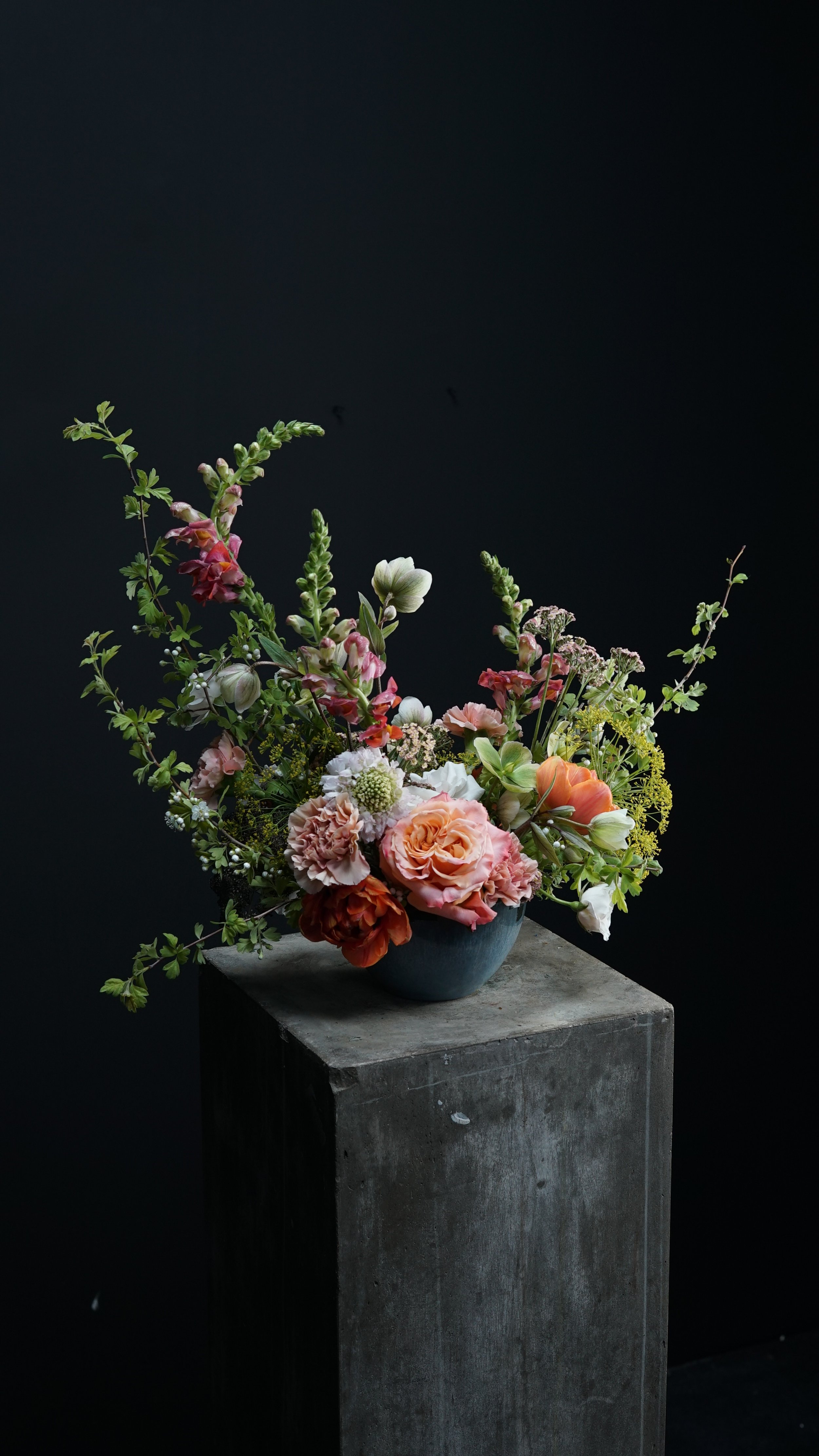 Jarrón con arreglo floral mixto en tonos pasteles, colocado sobre un pedestal de concreto, fondo oscuro.