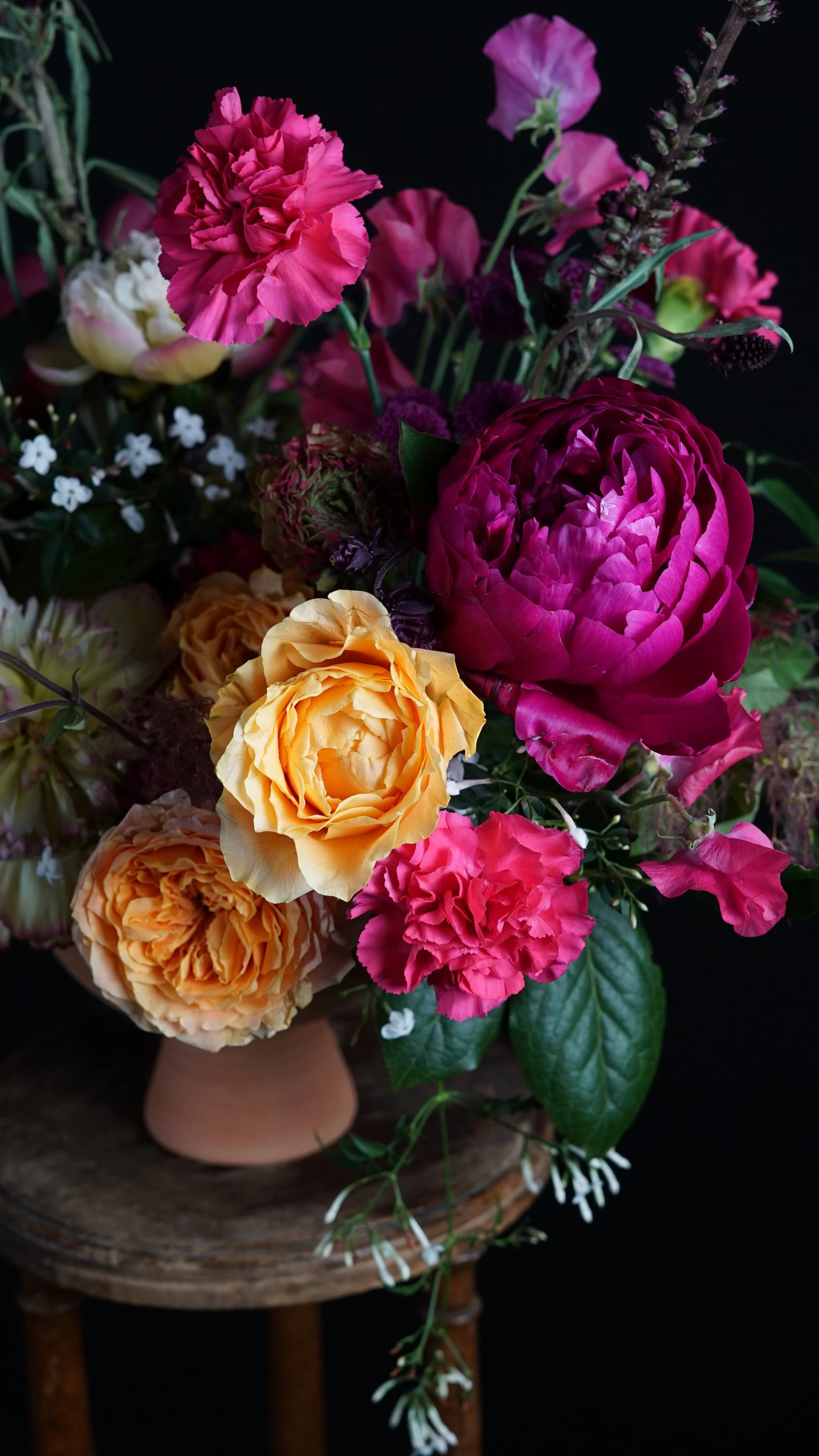 Ramo de flores variadas, incluyendo rosas amarillas, menta, y otras flores en tonos rosados, morados y blancos, en un jarrón de cerámica sobre una mesa de madera.