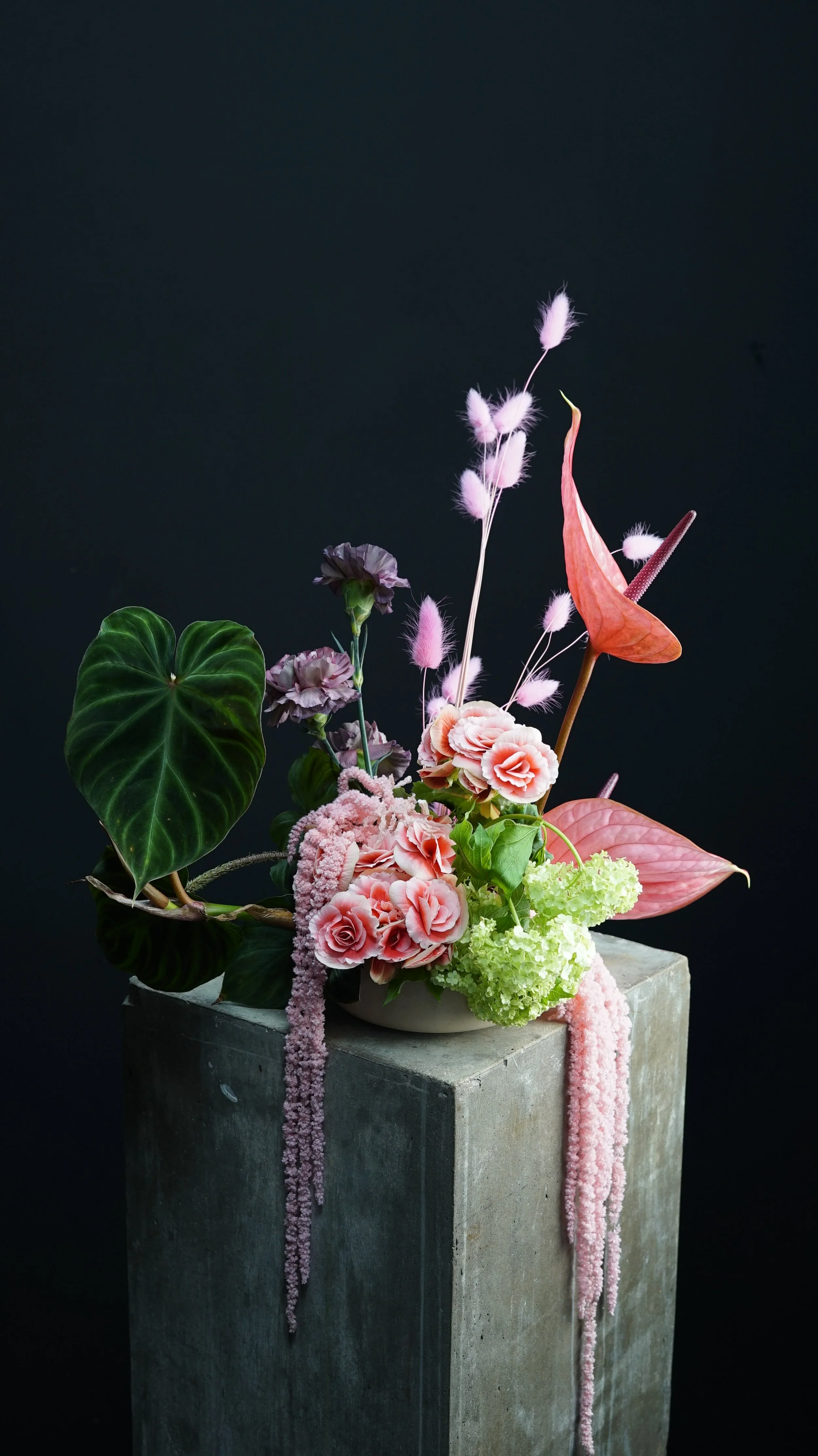 Arreglo floral con flores rosadas y verdes, hojas grandes, y cepillos rosados, colocado sobre una columna de cemento.