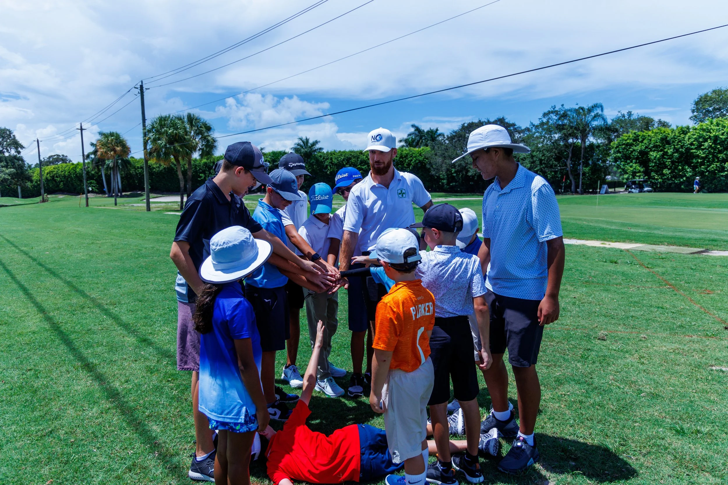Group of children and adults gathered on a golf course, with some children placing their hands together in the center, possibly for a team huddle or cheer. The setting is outdoors on a sunny day with blue skies and green trees.