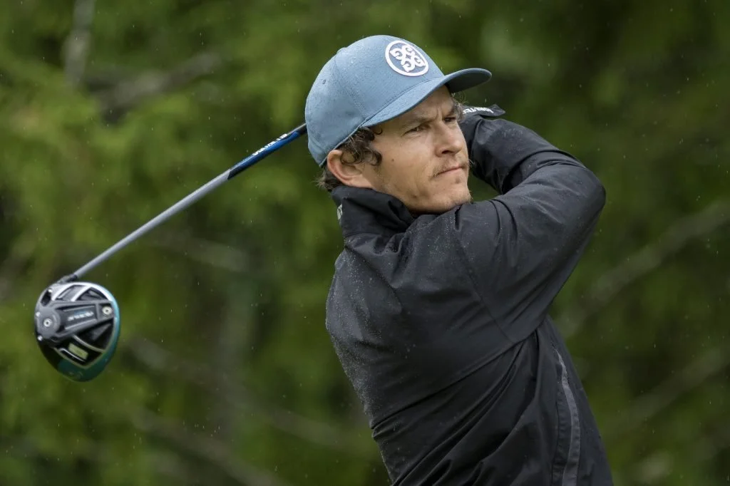A man playing golf, swinging a golf club outdoors with trees in the background.