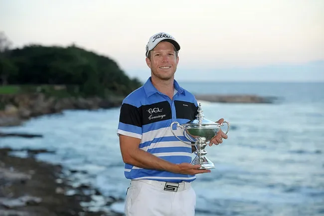 Male golfer holding a silver trophy on a beach with a body of water and greenery in the background.