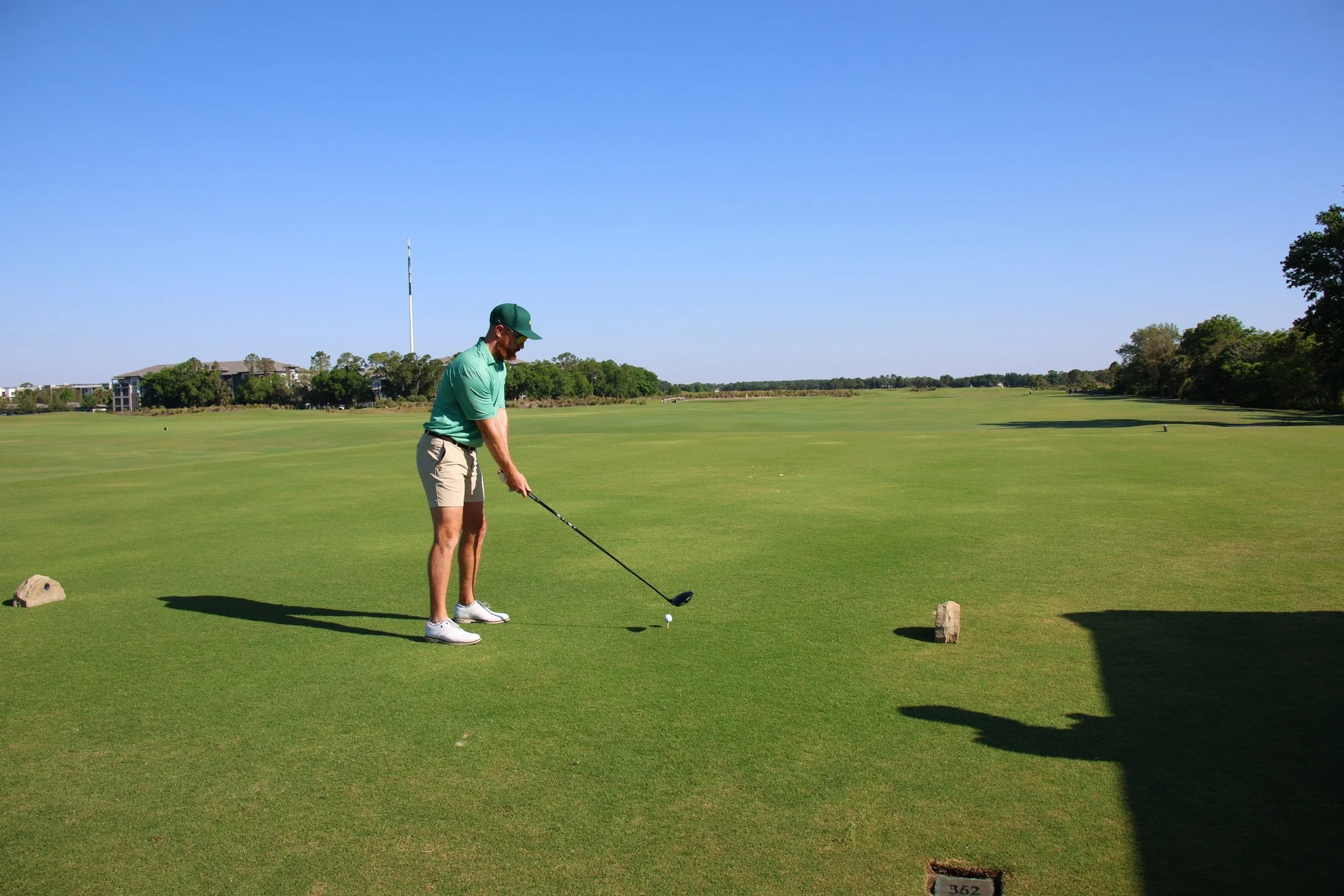 A man dressed in a green shirt, beige shorts, white shoes, and a green cap is about to hit a golf ball on a golf course under a clear blue sky. There are trees and buildings in the background.