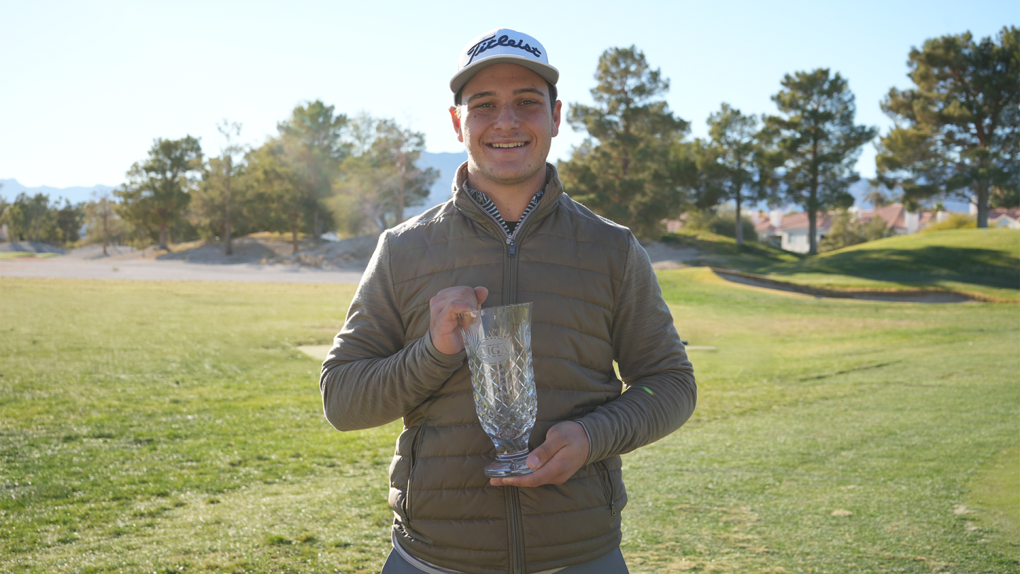 Young man smiling on a golf course holding a large glass trophy with trees and houses in the background.