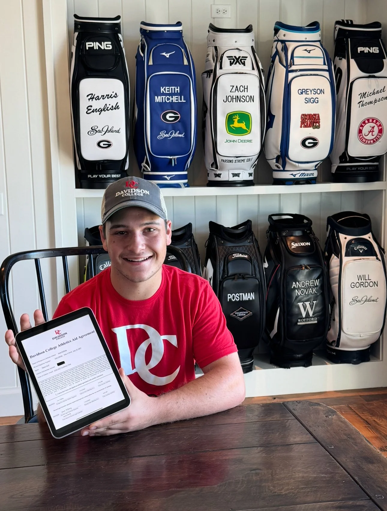 Young man smiling, wearing a gray baseball cap and red t-shirt, holding a tablet with a sports agreement document. Behind him, golf bags with personalized names and logos are displayed on a wall, and a wooden table is in front of him.