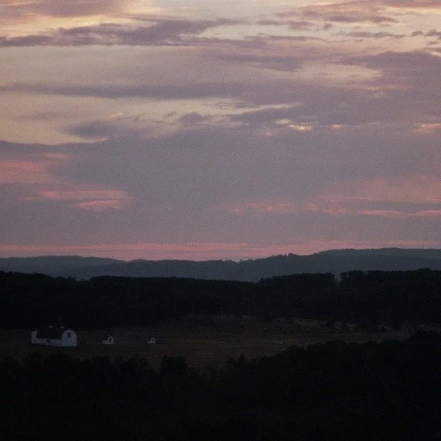 Sunset over rolling hills with a small white barn and farm buildings in the foreground, partly cloudy sky with pink and purple hues.