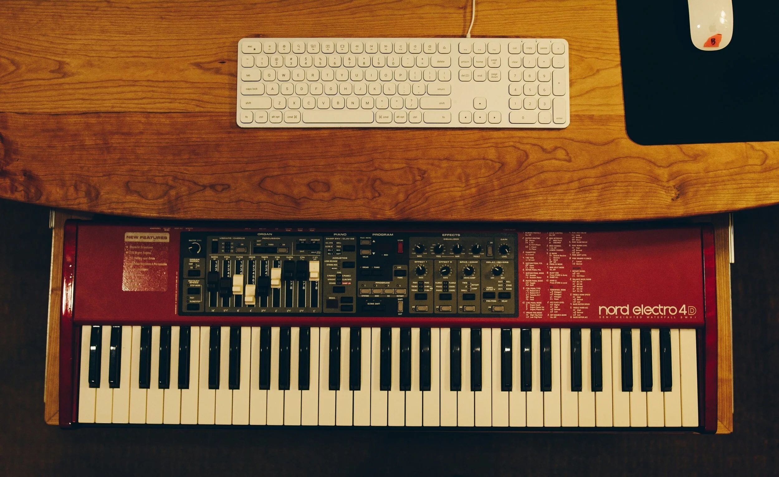 Top-down view of a wooden desk with a white computer keyboard, a black mouse on a black mouse pad, and a red Nord Electro 4D keyboard underneath.