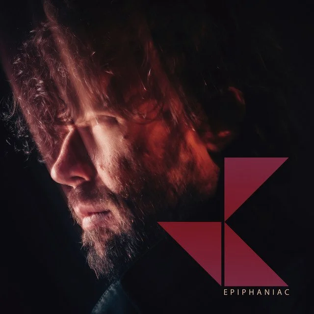 Close-up of a man's face with reddish-brown hair and beard, looking downward in soft lighting. The image features a logo with a stylized yellow letter 'K' and the text 'EPIPHANAC' at the bottom.