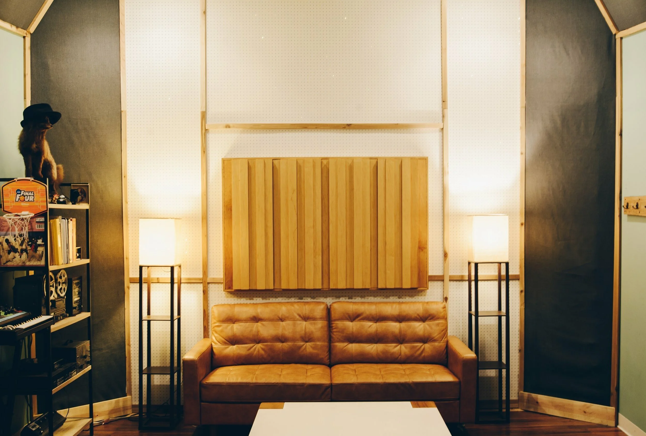 Living room with a brown leather sofa, two standing lamps on each side, a wooden wall panel behind the sofa, and a shelf with music equipment and a stuffed animal in the corner.