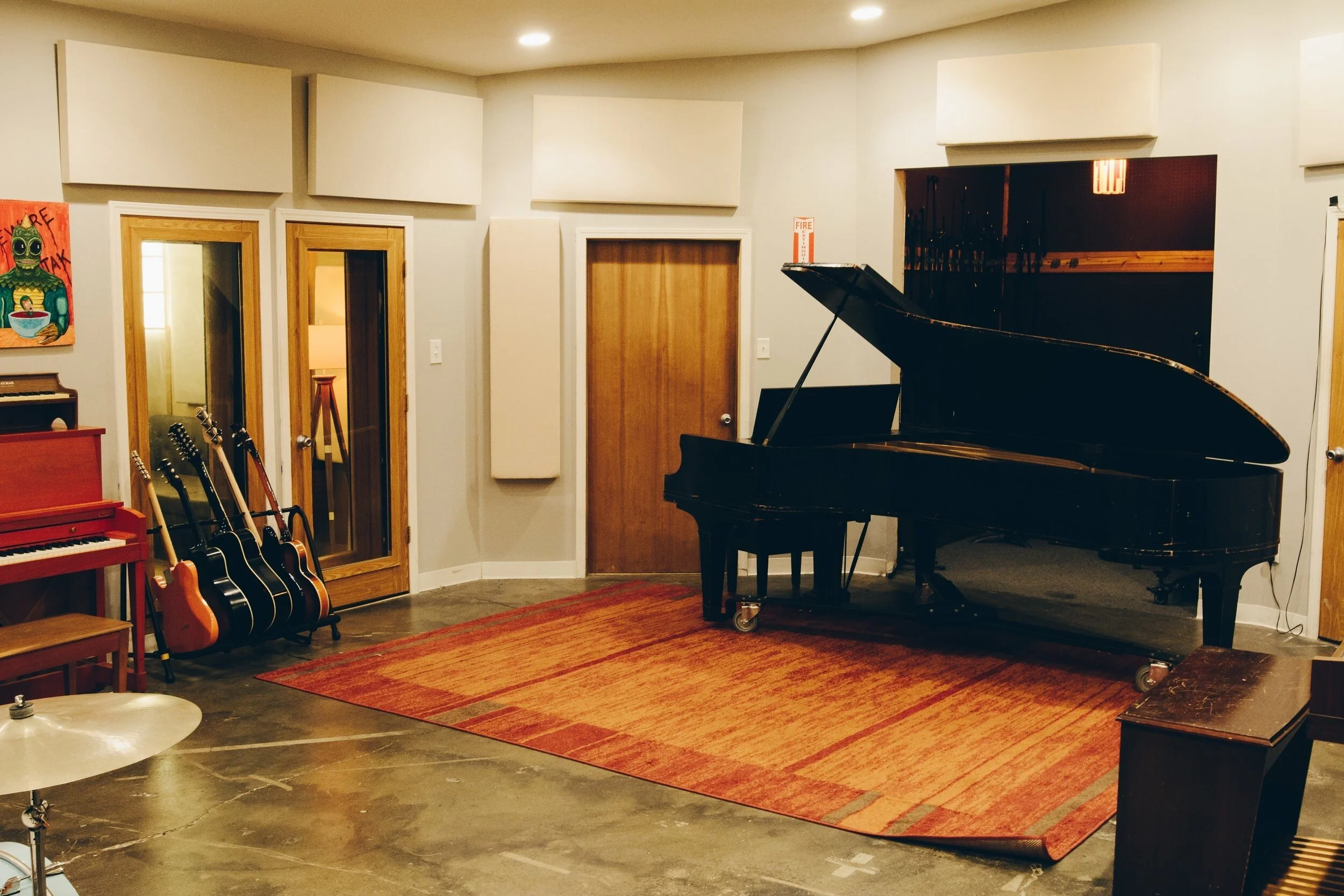 A music studio featuring a black grand piano, a red upright piano, several acoustic guitars, a drum cymbal, and a colorful painting on the wall.