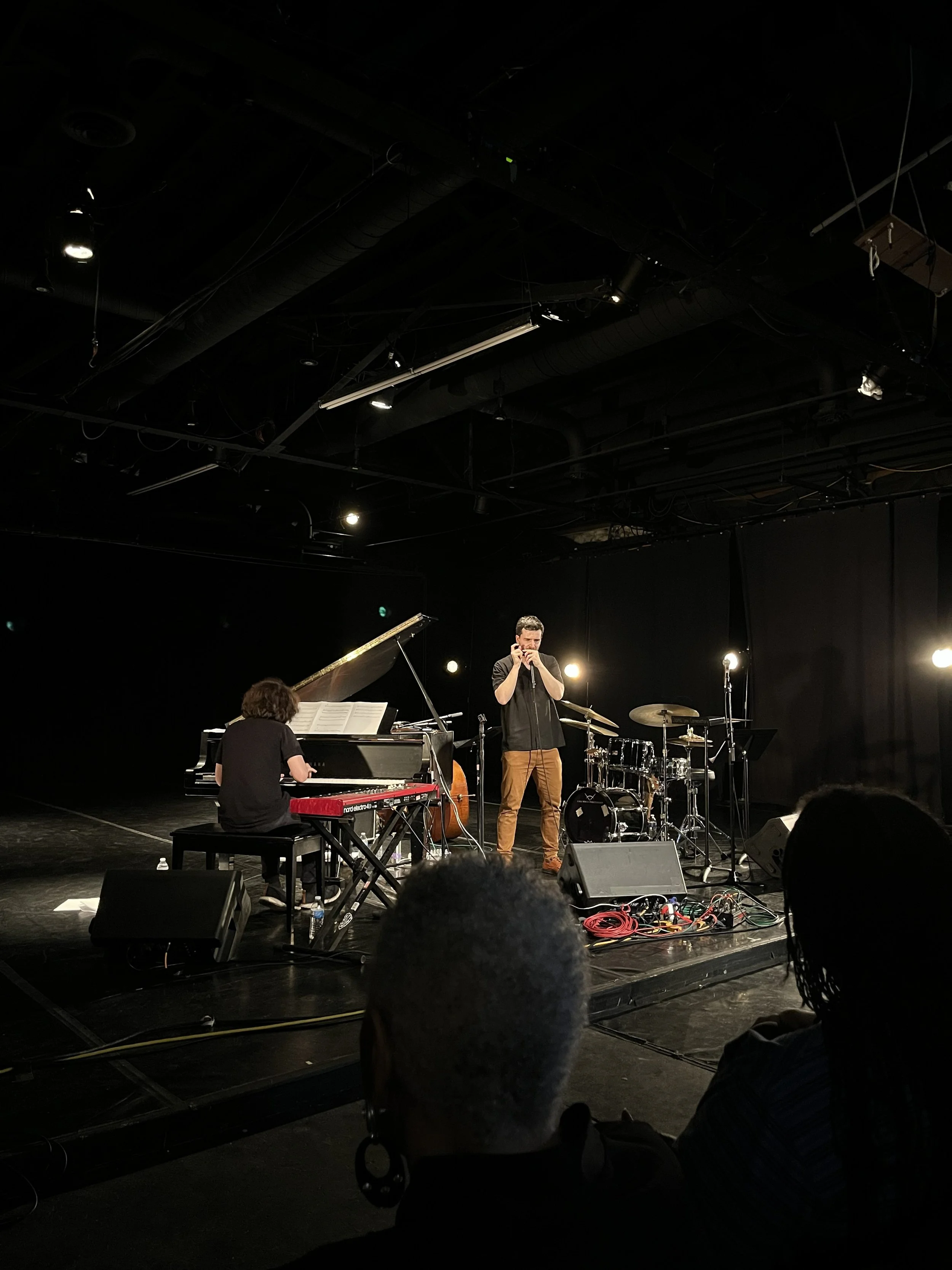 Musicians performing on a dark stage with a pianist, a drummer, and a singer, illuminated by stage lights, with audience members visible in the foreground.
