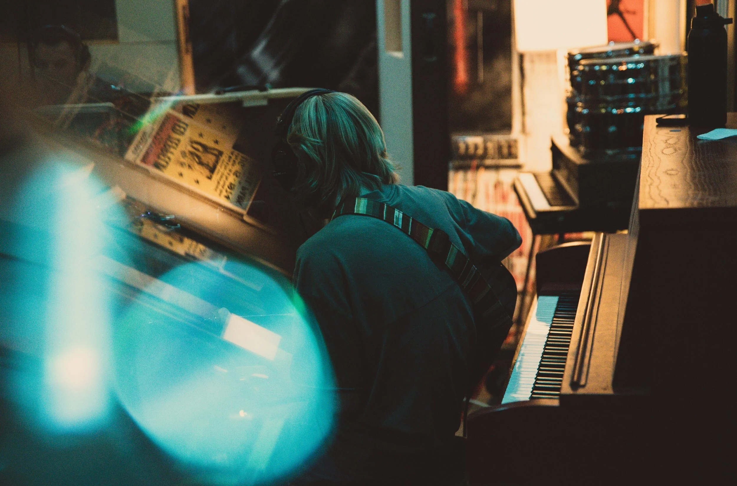 Person with head resting on a piano, wearing headphones, in a dimly lit room with music sheets and equipment.
