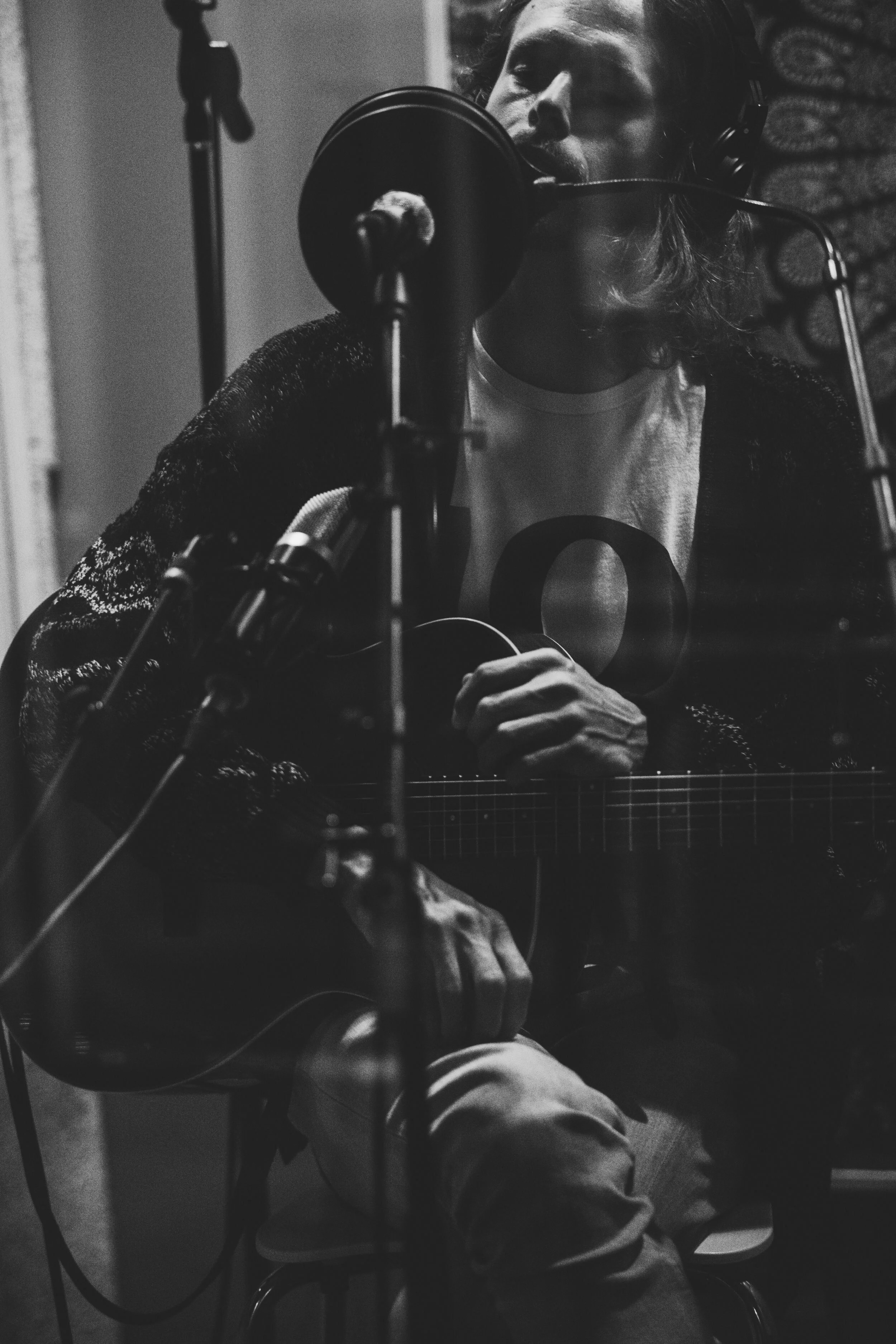 A man with long hair and a beard singing into a microphone while playing an acoustic guitar in a recording studio.