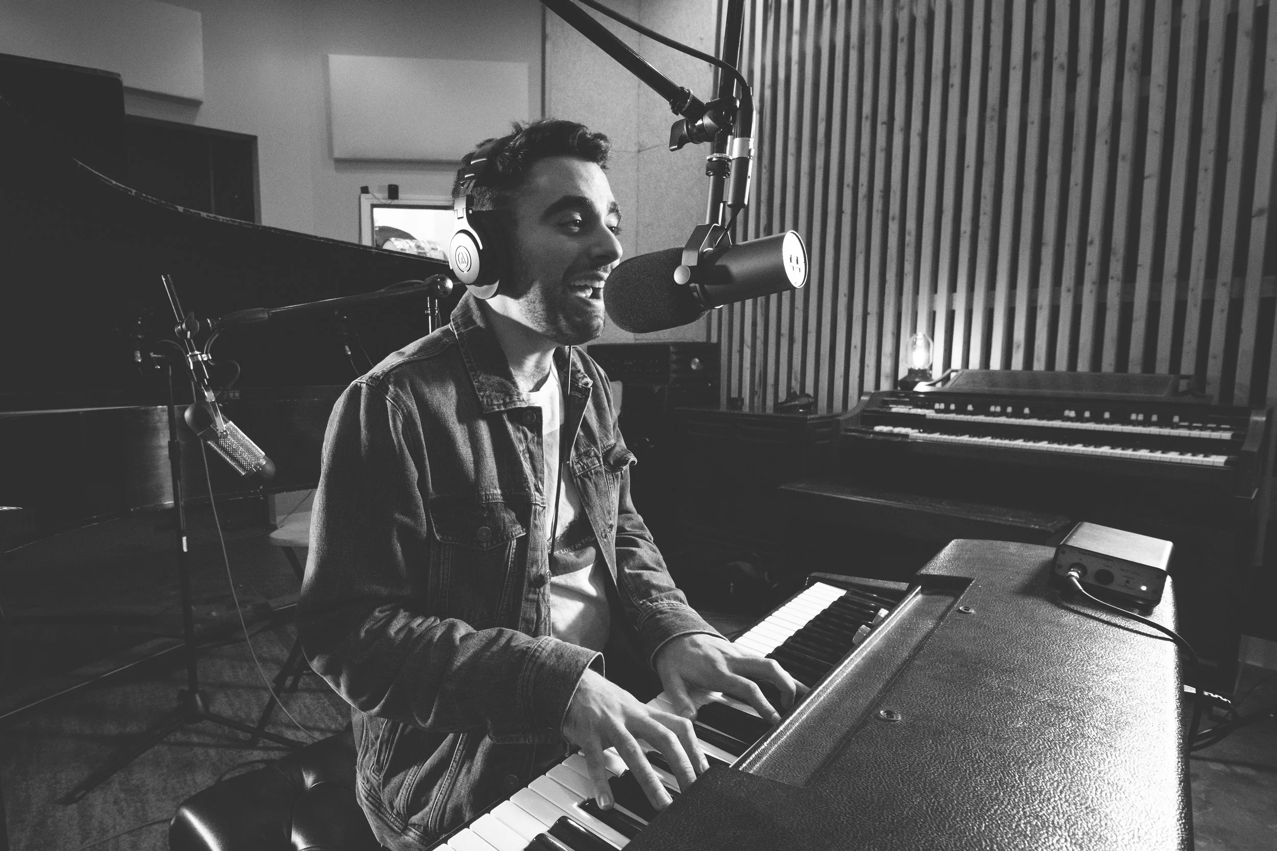Black and white photo of a man singing into a professional microphone while playing a keyboard in a recording studio, wearing headphones and a denim jacket.