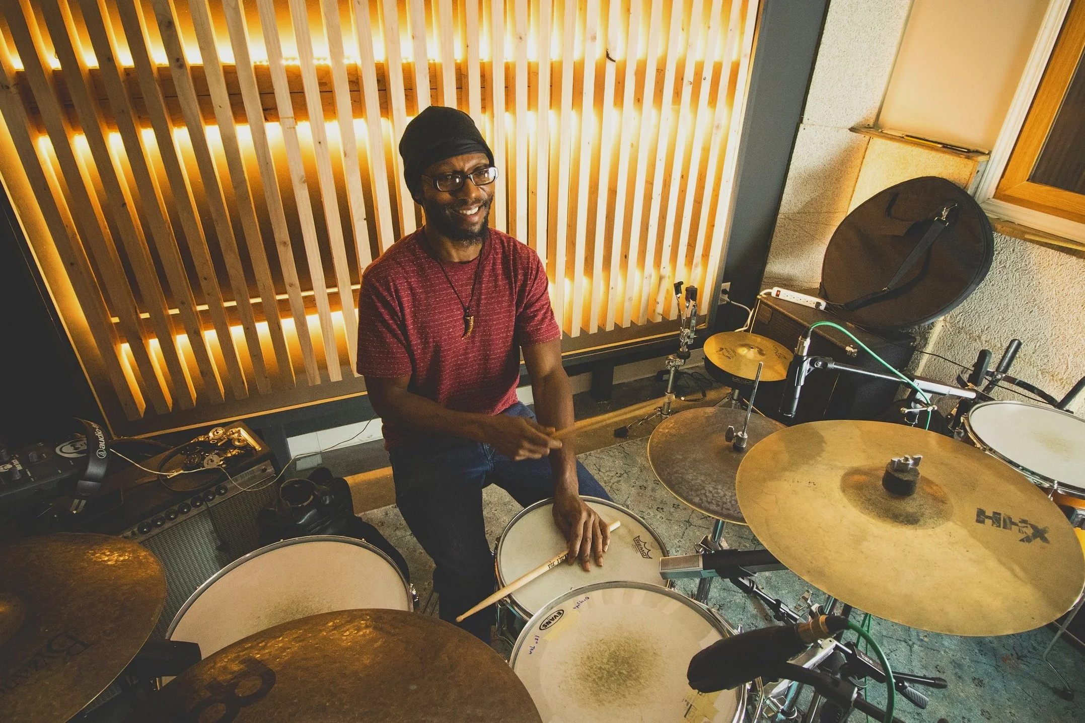 A man smiling while sitting behind a drum set in a music studio, wearing glasses, a black head covering, a red shirt, and a necklace, with wooden acoustic panels and equipment visible in the background.