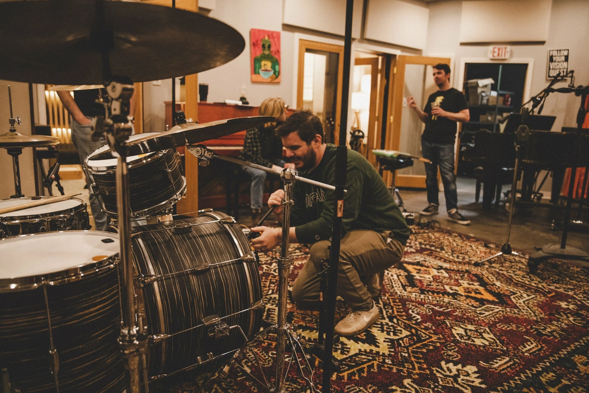 People in a music studio preparing musical instruments, with a man kneeling near a drum set, adjusting equipment, and others in the background in a cozy room with rugs and artwork.