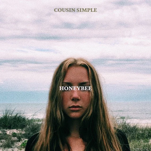 A young woman with long hair stands outdoors at the beach with grass and ocean in the background, and text overlays saying "COUSIN SIMPLE" at the top and "HONEYBEE" across her eyes.
