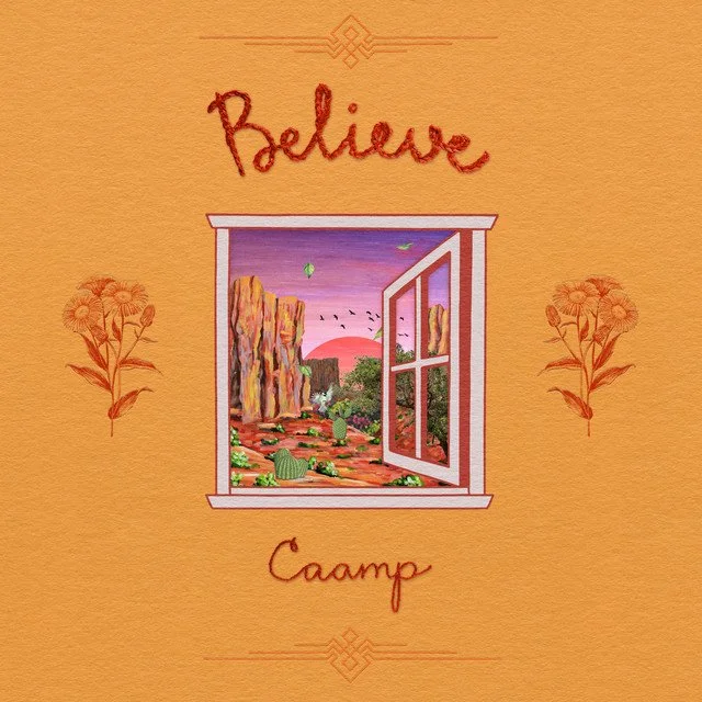 A decorative wall hanging with the word 'Believe' at the top and 'Camp' at the bottom, featuring an open window revealing a scenic landscape with cliffs, cacti, trees, and a pink sunset sky.