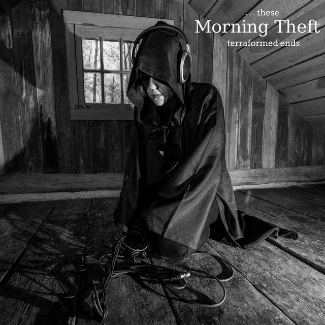 A person wearing a hooded cloak and headphones kneeling on a wooden floor in a rustic room, with tangled cables in front. The room has wooden walls and a small window. Text overlay says 'these Morning Theft terraformed ends.'