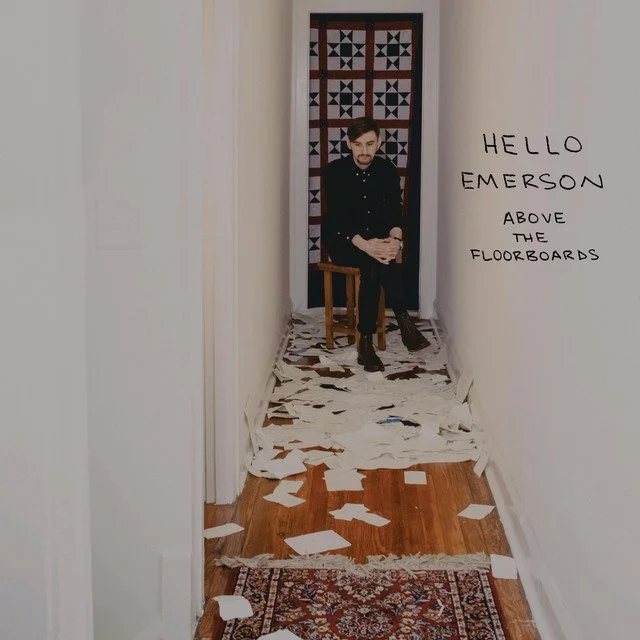 A man sitting on a wooden chair in a narrow hallway, with torn pieces of paper scattered on the floor. There is a large cutout of a person on the back wall and a handwritten note on the right wall that says, "HELLO EMERSON ABOVE THE FLOORBOARDS."