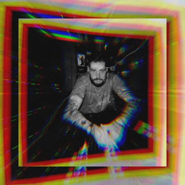 A person with glasses and tattoos leaning forward in a room with colorful, glitchy effects and a black background.