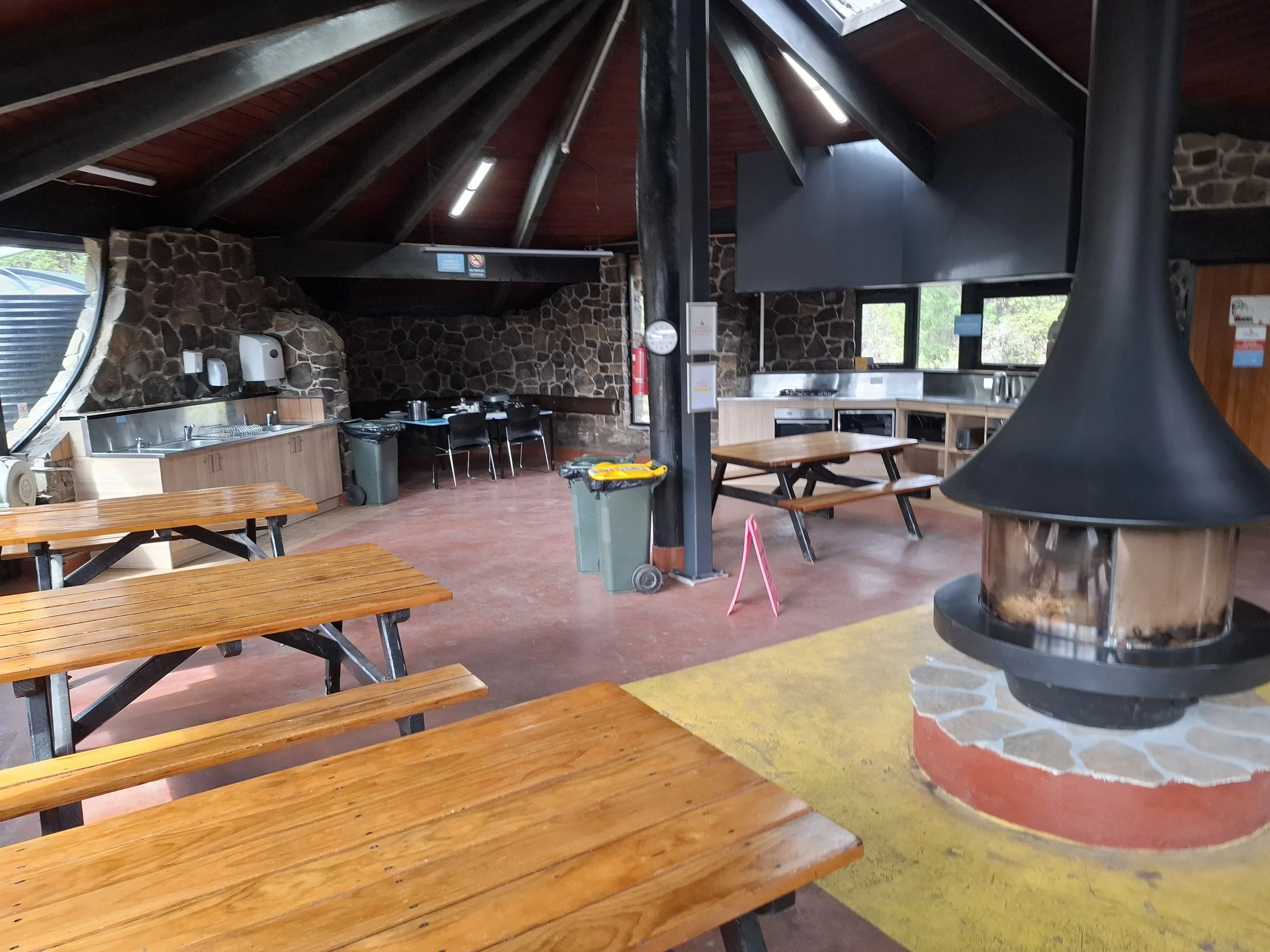 Indoor space with wooden picnic tables, stone wall, fire pit, soap and sanitizer dispensers, trash bins, large windows, and ceiling with wooden beams.