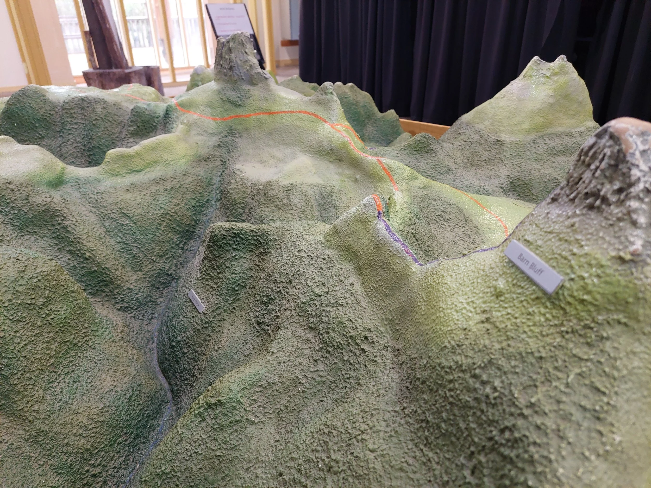 A scaled model of a mountainous landscape with labeled peaks, including Barn Bluff, with a textured green surface and painted trail lines, displayed indoors.