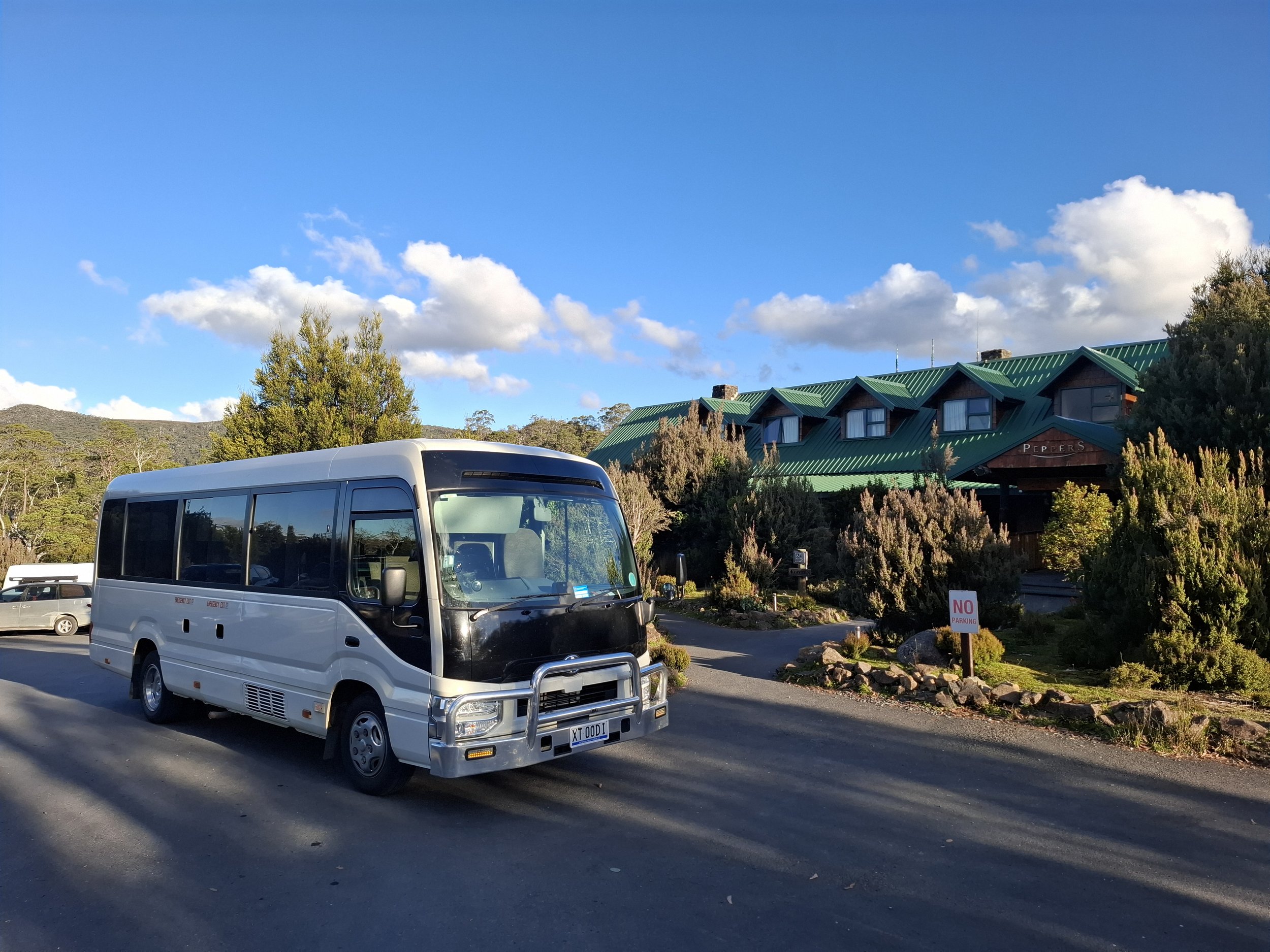 A white shuttle bus parked in a parking lot with a building with a green roof and trees in the background, under a blue sky with scattered clouds.