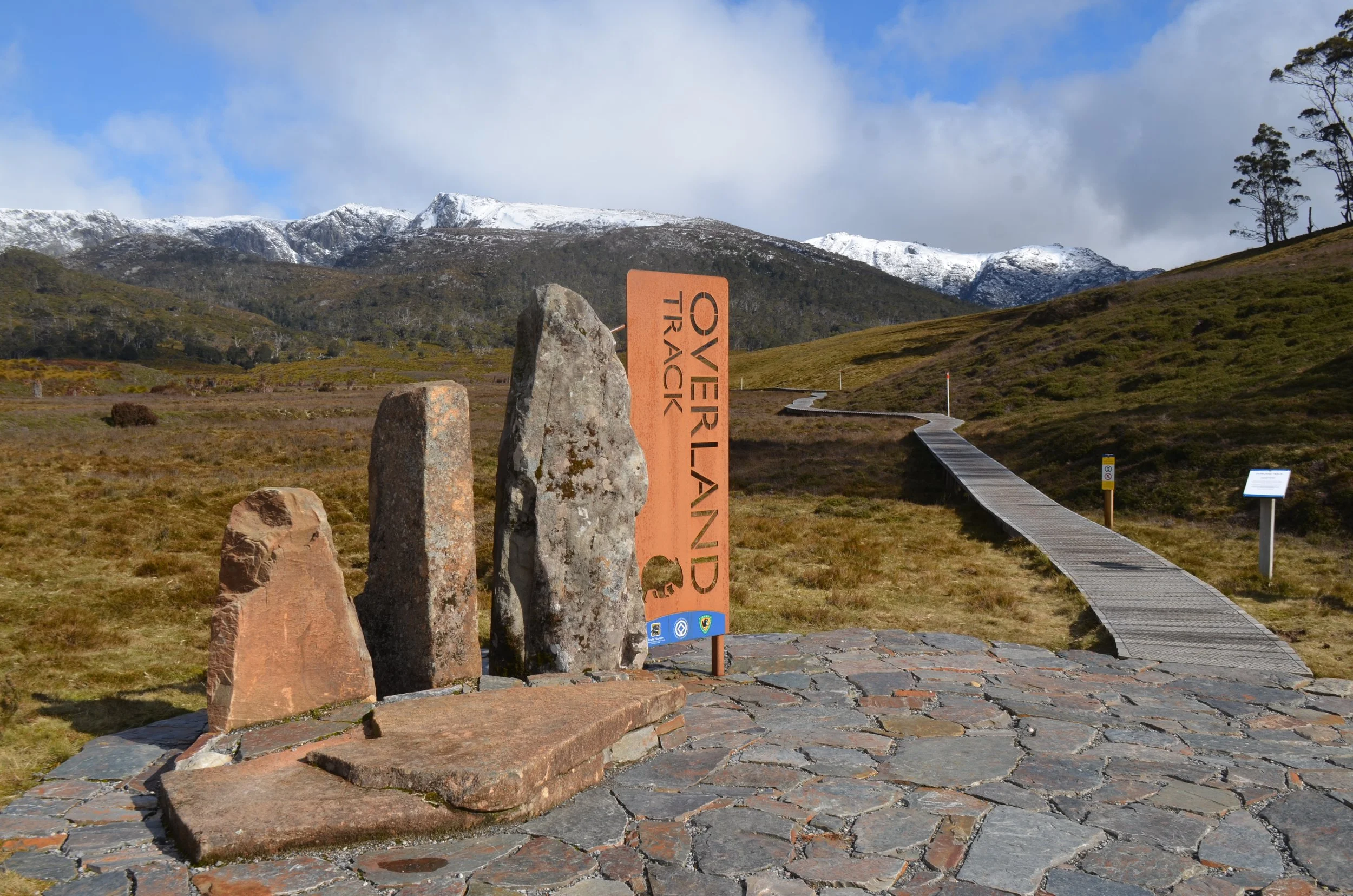 A scenic view of a mountainous landscape with snow-capped peaks, a winding boardwalk through grassy terrain, and a sign indicating 'Overlander Track' with a small bear symbol at the bottom.