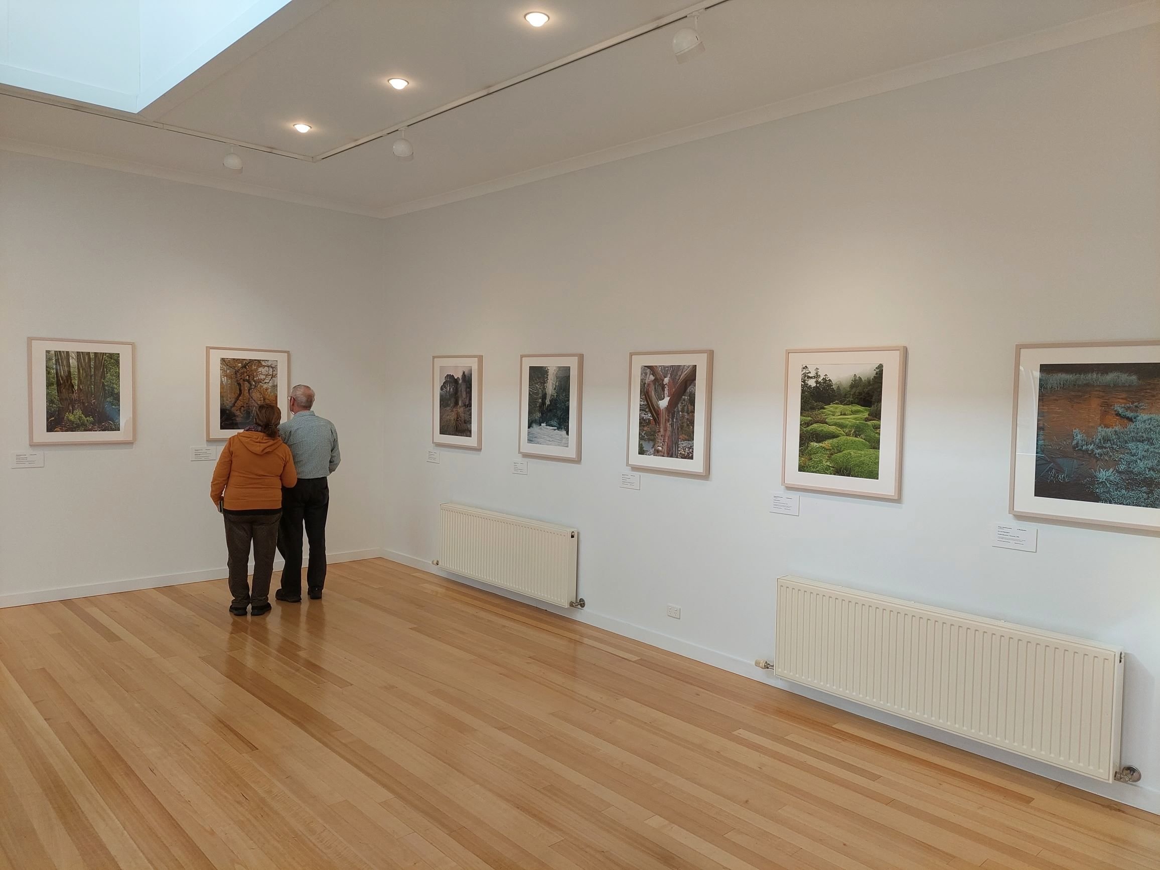 Two people, a man and a woman, are standing and looking at framed photographs of trees and landscapes displayed on a white gallery wall. The gallery has bright lighting, a wooden floor, and a white radiator below the photos.