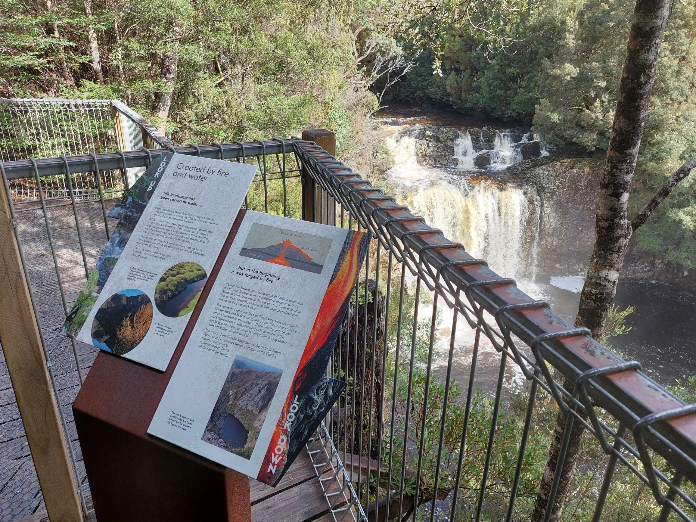 A view of a waterfall cascading over rocks surrounded by trees in a lush forest, taken from a viewing platform with informational signs.