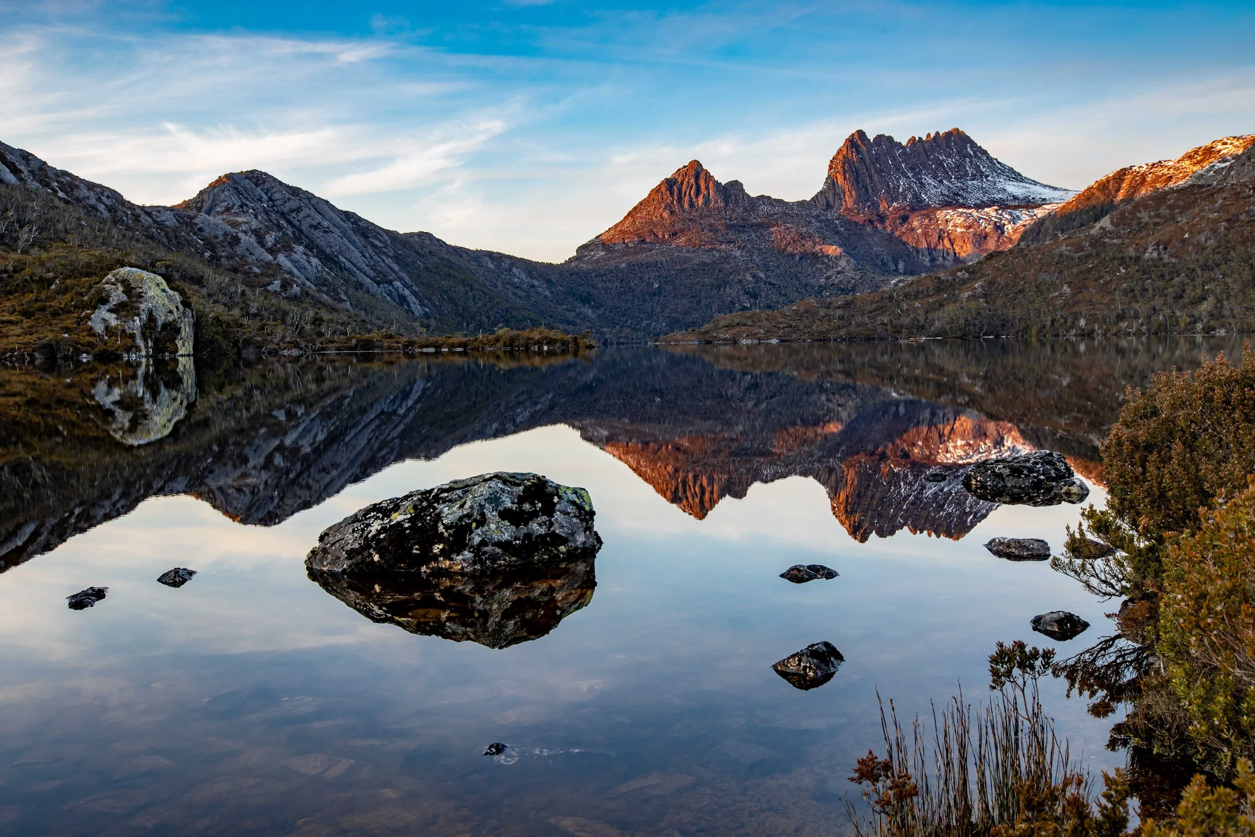 Reflection of mountain peaks and blue sky with some clouds on a calm lake with rocks and shrubs in the foreground.