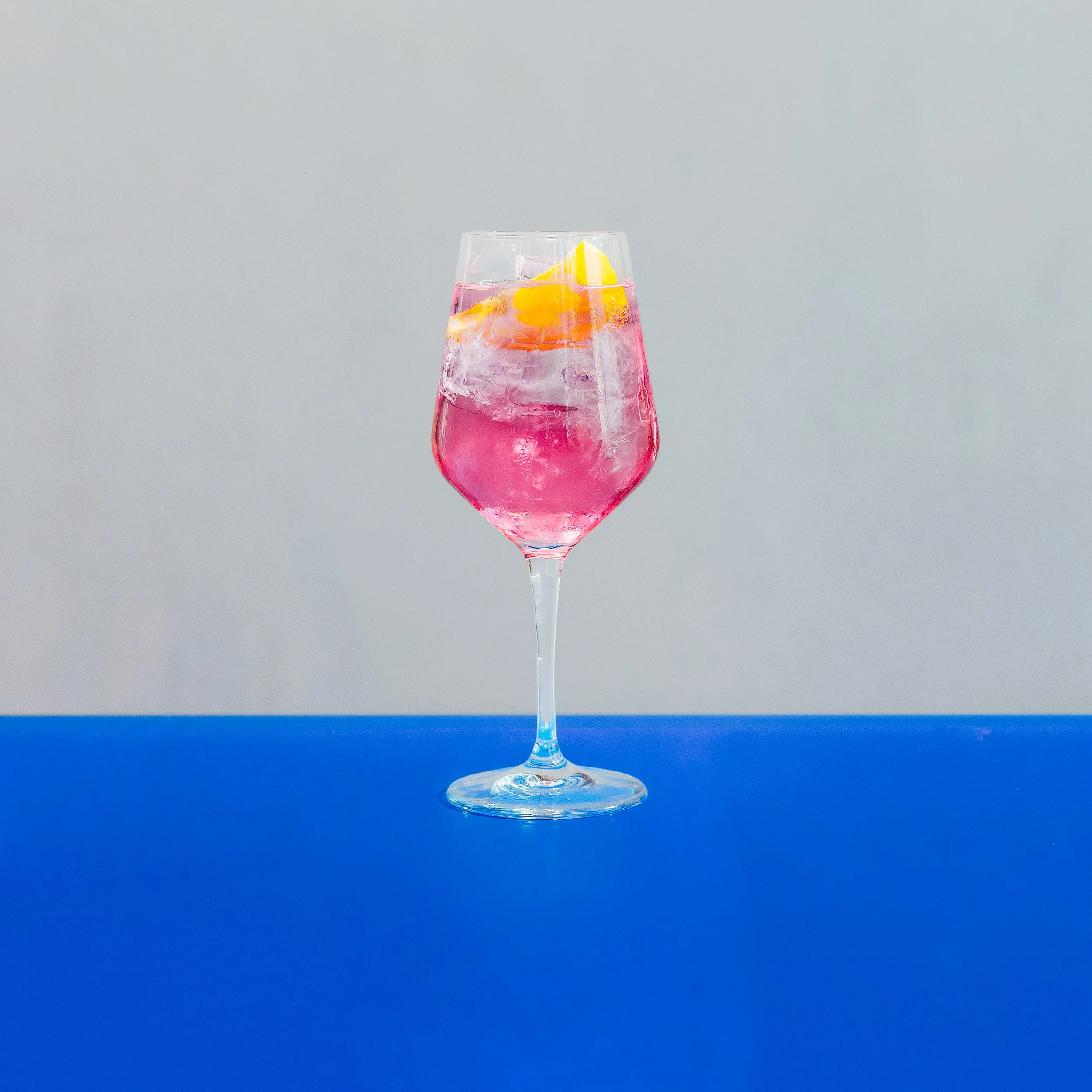 A pink Sorta Rosa spritz with ice and a blood orange wedge in a wine glass on a bright blue table