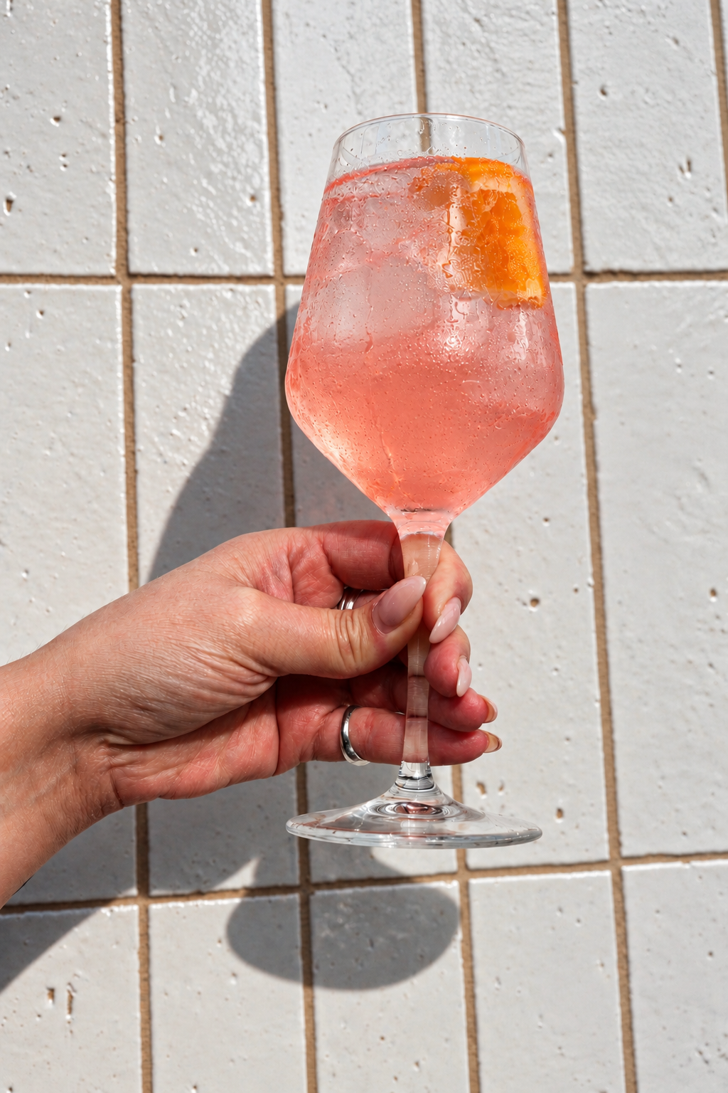 A hand holding up a pink Sorta Rosa spritz with an orange slice against a sunlit white tiled wall