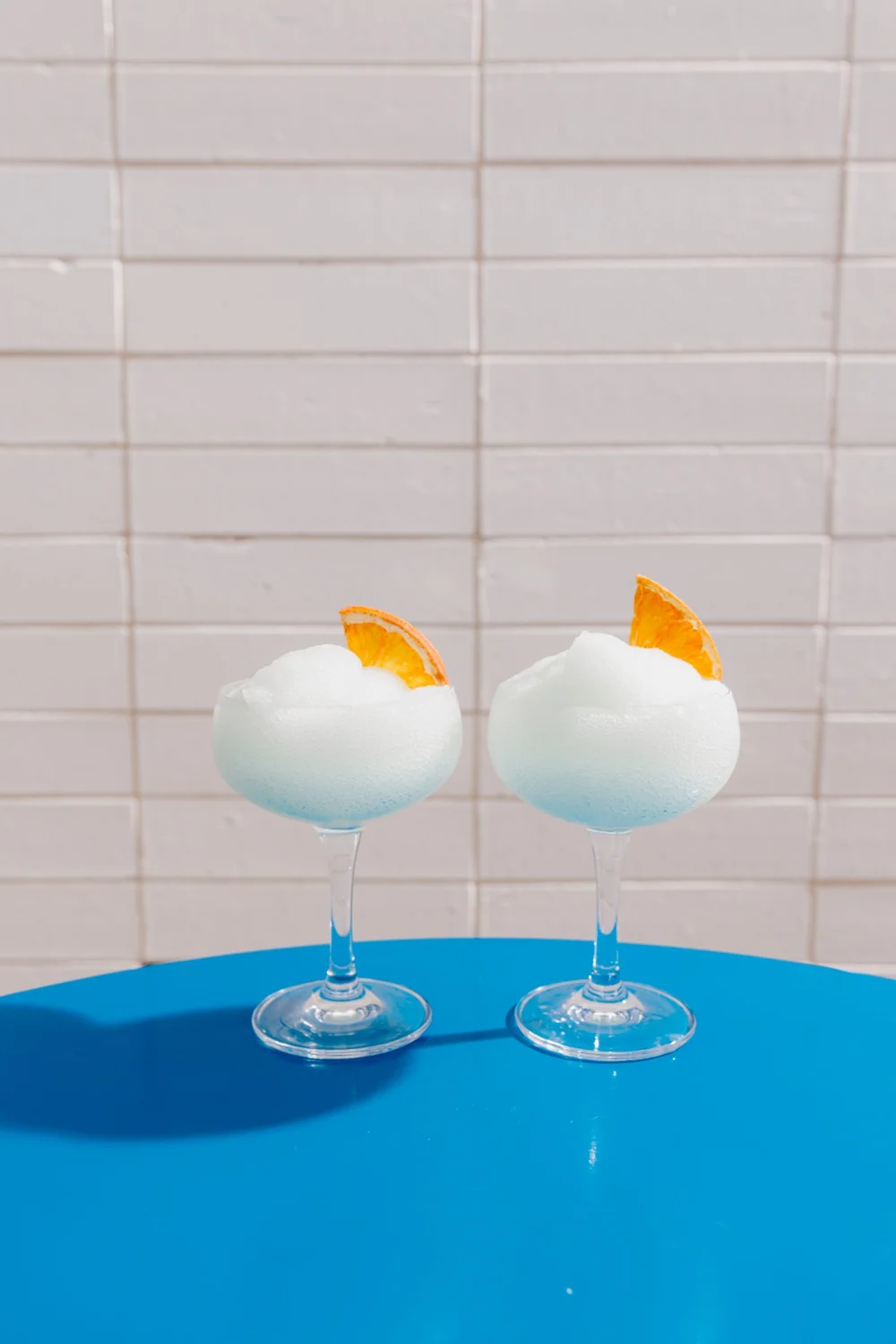 Two white frozen cocktails with dried orange garnish on a bright blue table at Good Heavens Rooftop Bar. Frozens $19 each, $10 on Sundays and $12 in happy hour, weekdays 3-6pm.