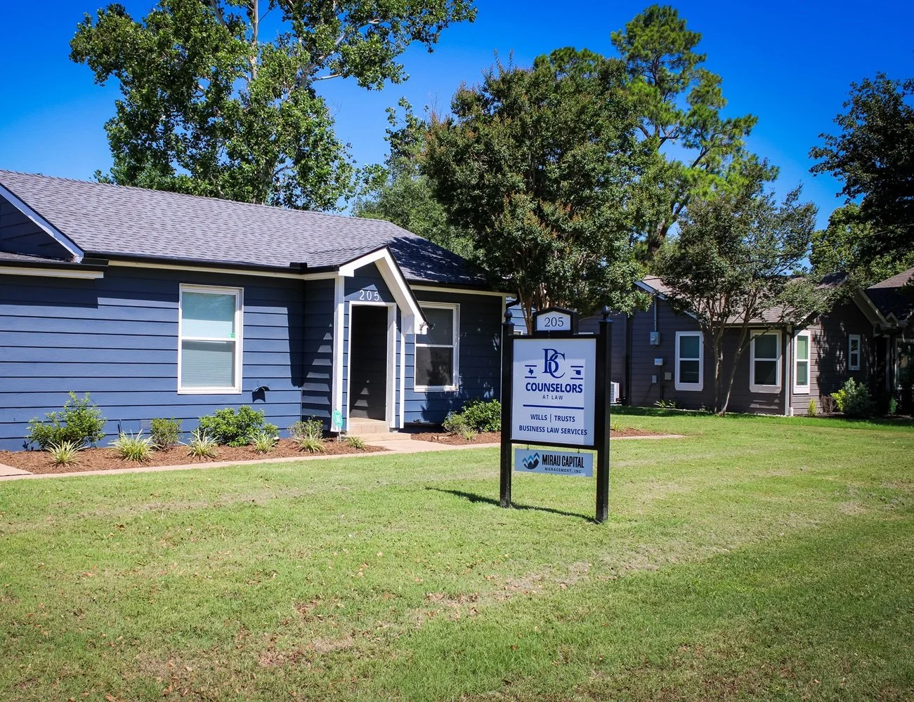 Exterior of a blue house with a sign reading 'Counselors at Law, Wills, Trusts, Business Law Services' on a grassy lawn.