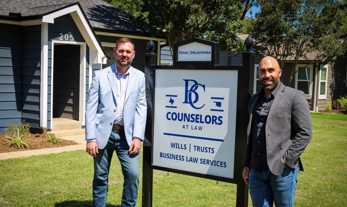 Two men standing outside a law office with a sign reading 'Counselors at Law - Wills, Trusts, Business Law Services' from Texas and Oklahoma.