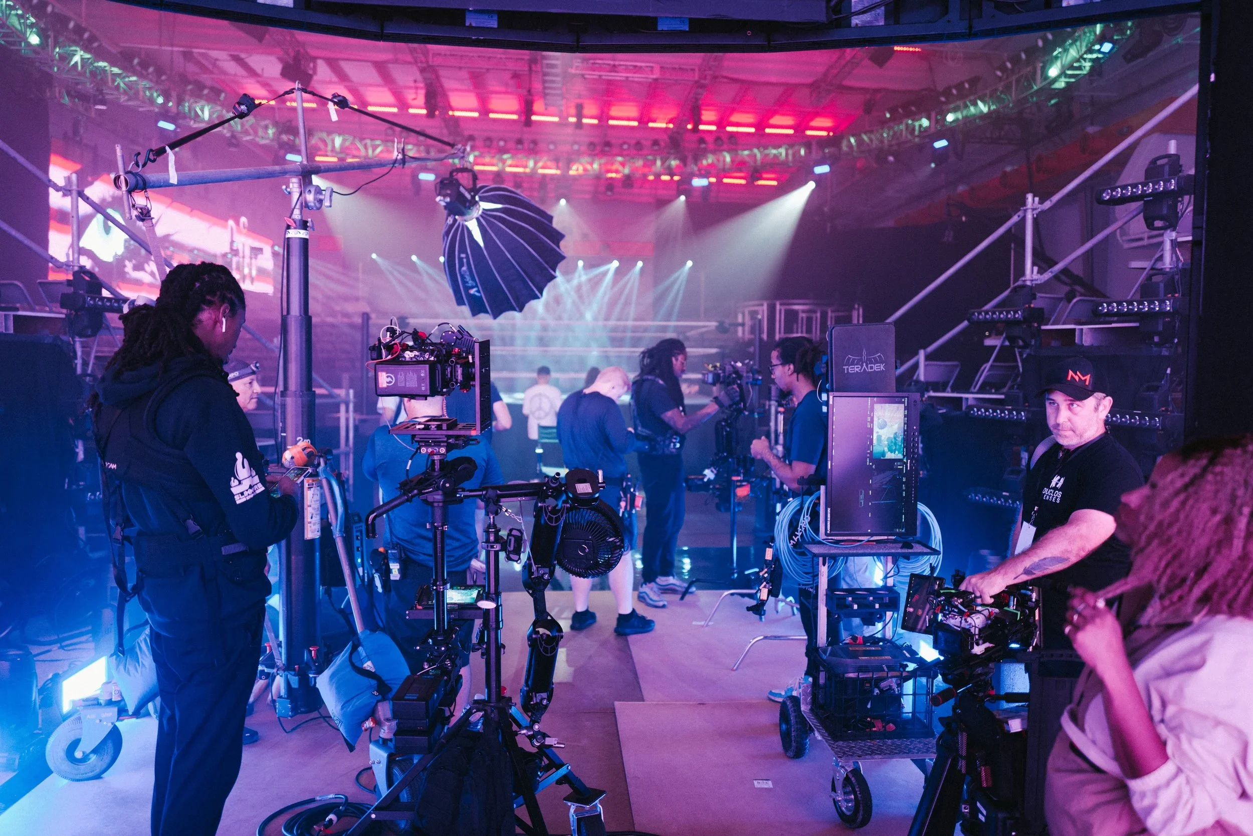 Filming crew setting up equipment on a stage with colorful lighting and audience seating area visible in the background. Libertine Studios - Joshua Libertine