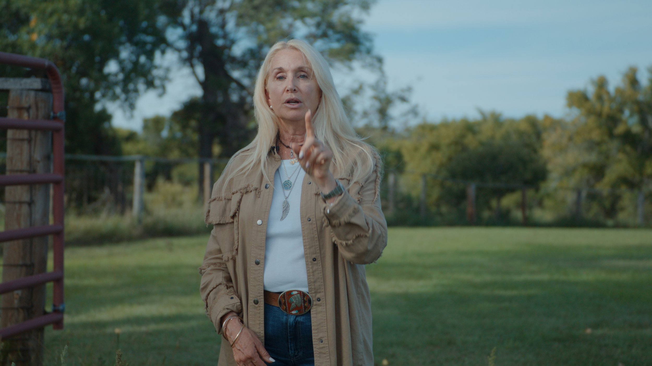 A woman with long blonde hair standing outdoors in front of trees, wearing a beige jacket over a white shirt, with jewelry and a large belt buckle, holding up her index finger while speaking.