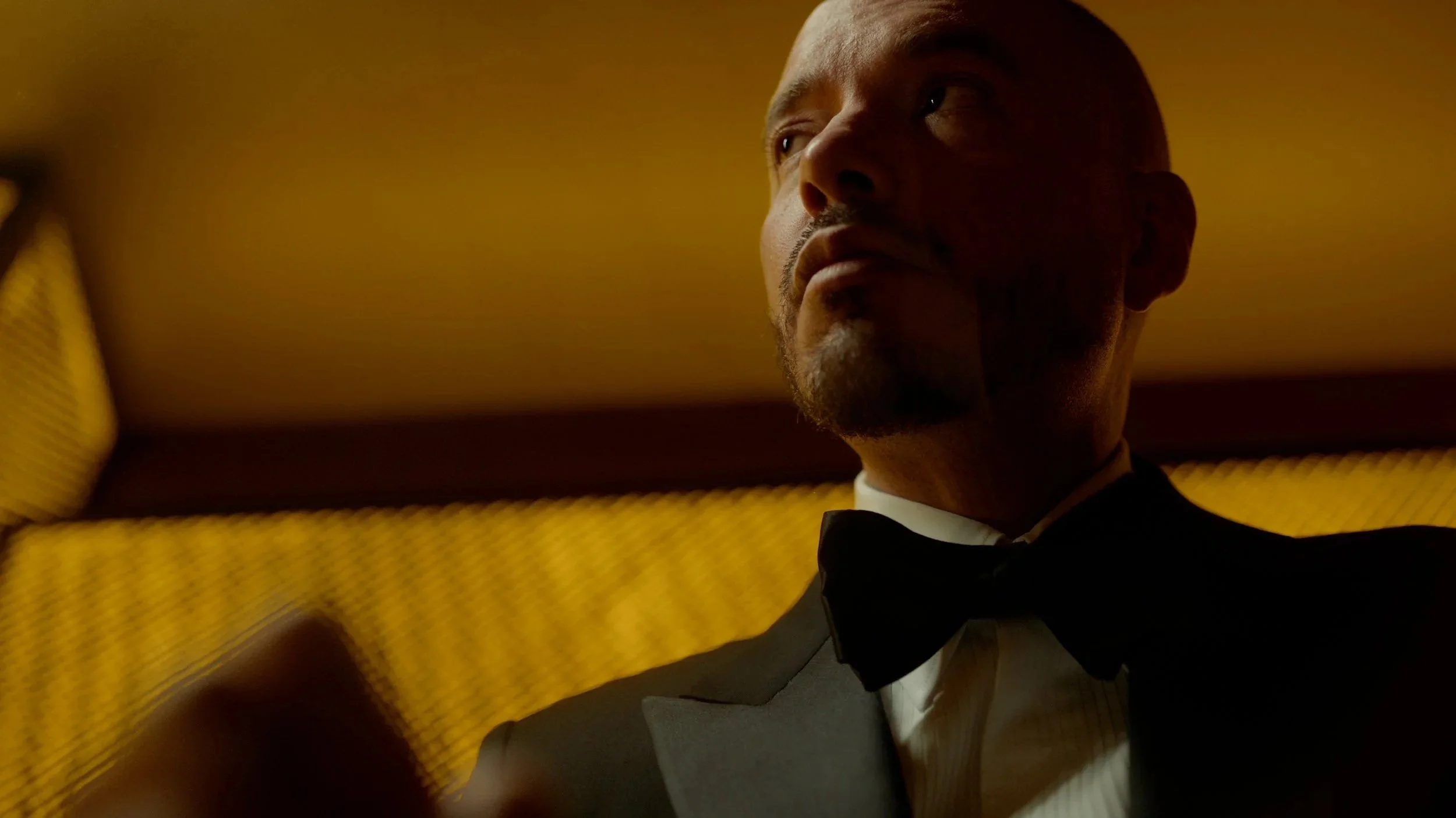 A man J. Balvin in a tuxedo with a bow tie is looking to the side in a dimly lit room with yellow and brown decor.