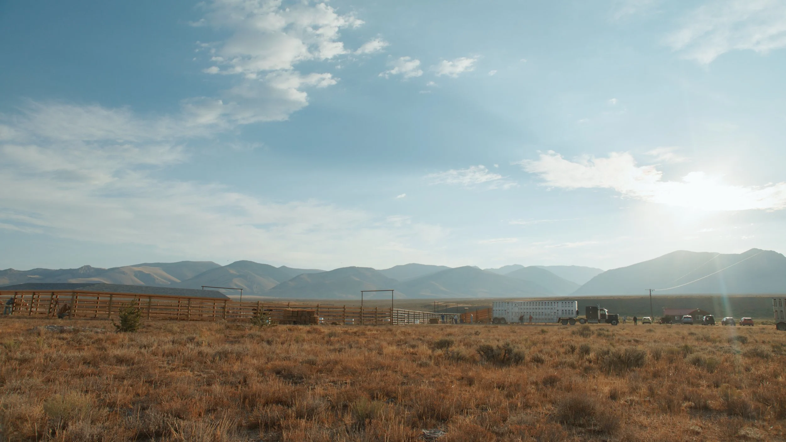 A vast open landscape with dry grass in the foreground, a fenced area with some trees, and mountains in the distance under a partly cloudy sky, with the sun shining from the right.